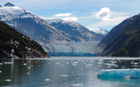 Calm water with iceberg bits in front of Dawes Glacier being framed by mountains on a sunny day in Alaska.