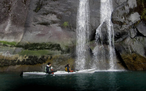 Two kayakers paddling past twin waterfalls flowing down high rock walls in Alaska.
