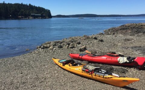 1 yellow & 1 red kayak sit on a shore of pebbles beside calm blue water with tree-lined shores in the background on a sunny day during the Voyage Through the San Juan Islands small ship cruise.