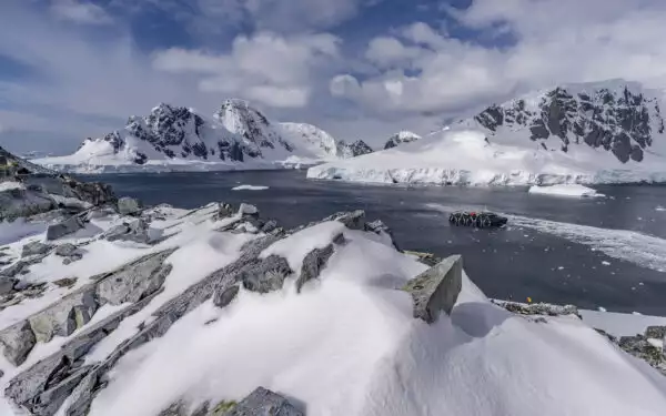 A luxury Antarctica cruise ship with a blue hull seen among mountains and snow with hikers on land