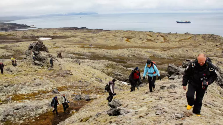 Small group of Arctic travelers dressed warmly in various colors hikes up beige, rocky tundra as a small ship sits offshore.