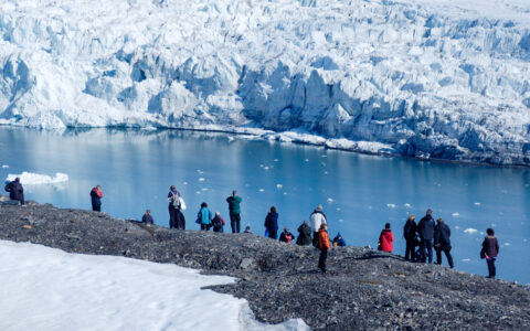 Small group of Arctic Ocean Voyage travelers stand in the sun atop a rocky overlook to view fjord & white, jagged glacier below.