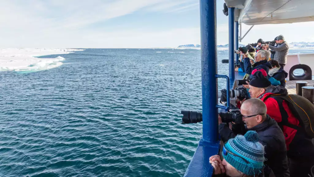 Group of Arctic Ocean Voyage travelers stand on deck of small ship & photograph ice edge over railing on a sunny day.