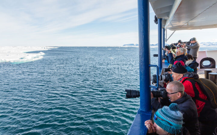 Group of Arctic Ocean Voyage travelers stand on deck of small ship & photograph ice edge over railing on a sunny day.