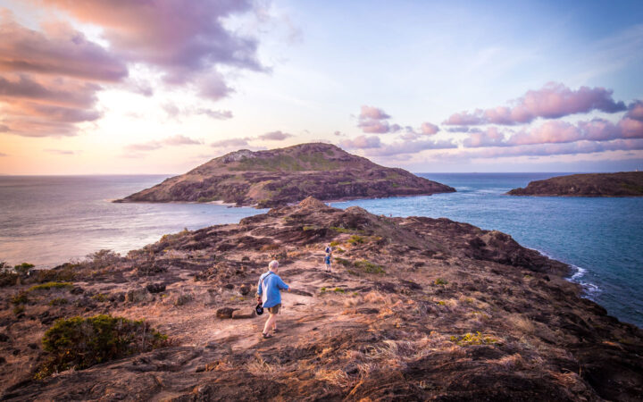 Australia travelers hike along the rocky, sandy top of an island at sunset with purple clouds above & blue sea below.