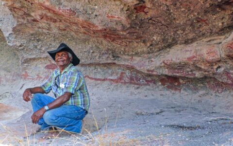 Man in dark cowboy hat, checkered shirt & jeans kneels down & smiles beneath indigenous rock art, seen in Arnhem Land.