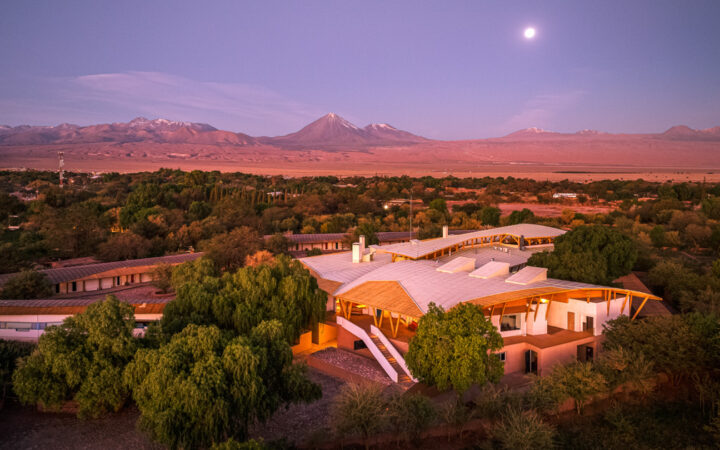 Aerial view of Explora Atacama Lodge in Chile at sunset, with white & wood construction among green trees & nearby desert.