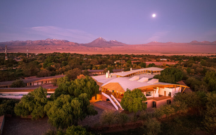 Aerial view of Explora Atacama Lodge in Chile at sunset, with white & wood construction among green trees & nearby desert.
