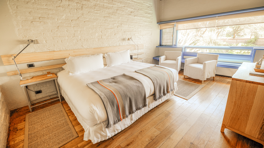 Yali Room at Explora Hotel Atacama with king bed in white linens, small wood table, large window, white walls & blue accents.
