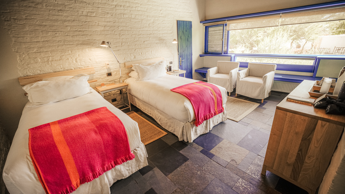 2 twin beds in white & pink with beside table, wood dresser, 2 chairs & view window in Tulur Room at Explora Atacama Lodge in Chile.