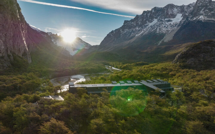 Aerial view of a dark metal, rectangular hotel sitting beside a river in a valley of trees by tall, snow-capped peaks.
