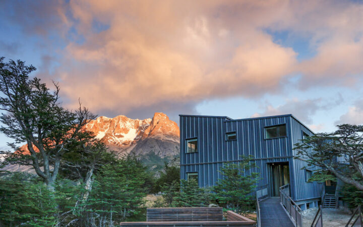 Modern blue metal building sits beside a wooden walkway & trees with alpenglow on the snow-covered peaks just beyond.