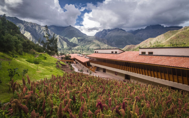 Looking down on the long, stone & wood Explora Sacred Valley lodge in a bright green Andean valley with purple flowers.