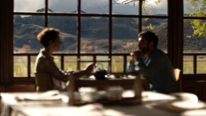 Man & woman dine at a 2-top table beside large window with wood lattice & views to green fields & mountains in Patagonia.