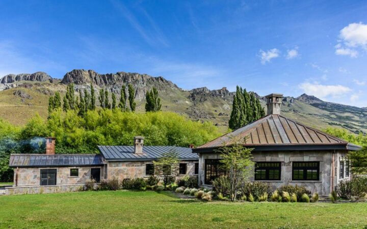 Exterior of stone building with 3 large interconnecting pieces sitting among grass & rugged peaks under a blue sky in Patagonia.