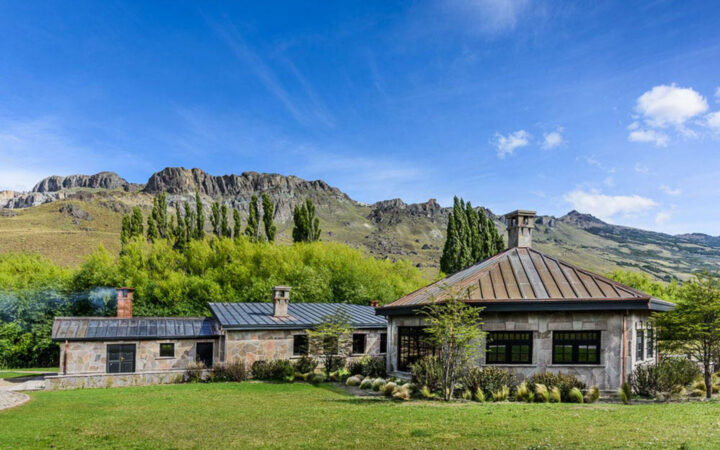 Exterior of stone building with 3 large interconnecting pieces sitting among grass & rugged peaks under a blue sky in Patagonia.