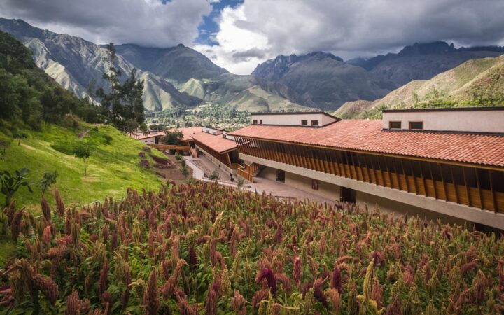 Looking down on the long, stone & wood Explora Sacred Valley lodge in a bright green Andean valley with purple flowers.