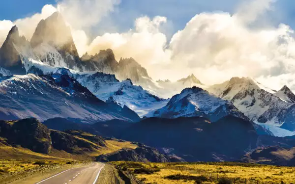 A road extends into Patagonia mountains covered in light snow and clouds with the sunlight behind them