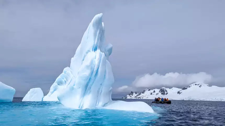 beautiful iceberg glowing with white and blue on a cloudy day next to a zodiac full of explorers
