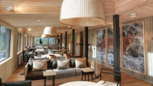 Common area at Hotel Explora El Chalten with light wood, panoramic windows, couches & coffee tables under woven lamps & wildlife art.
