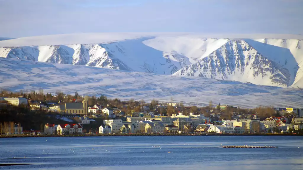 Seaside town of Akureyri, Iceland in winter with beige & white buildings backed by tall, snowy mountains.
