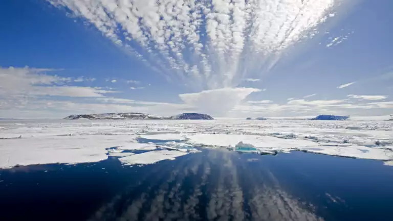 Edge of pack ice in large & small chunks with cloud reflections on glassy water during a sunny day near Iceland.