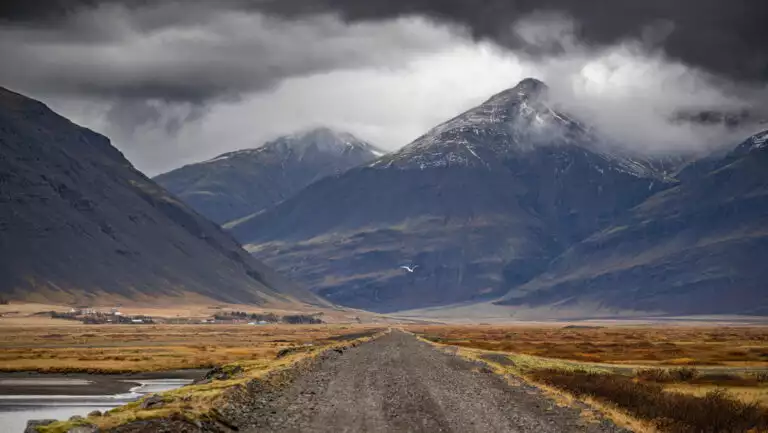 Secluded gravel road in Iceland leads past autumnal flatlands to tall volcanic mountains in the clouds.