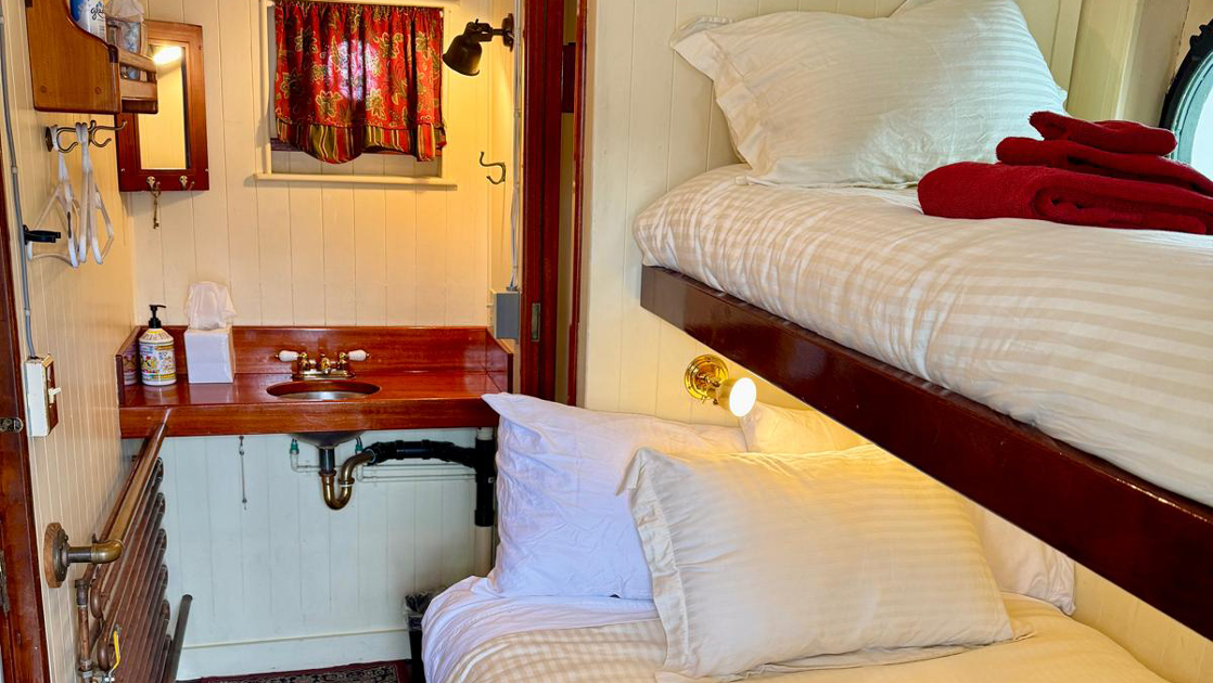 Cabin 1 on mv Catalyst with wooden bunk beds in white linens with red towels, porthole, white walls & bathroom sink.