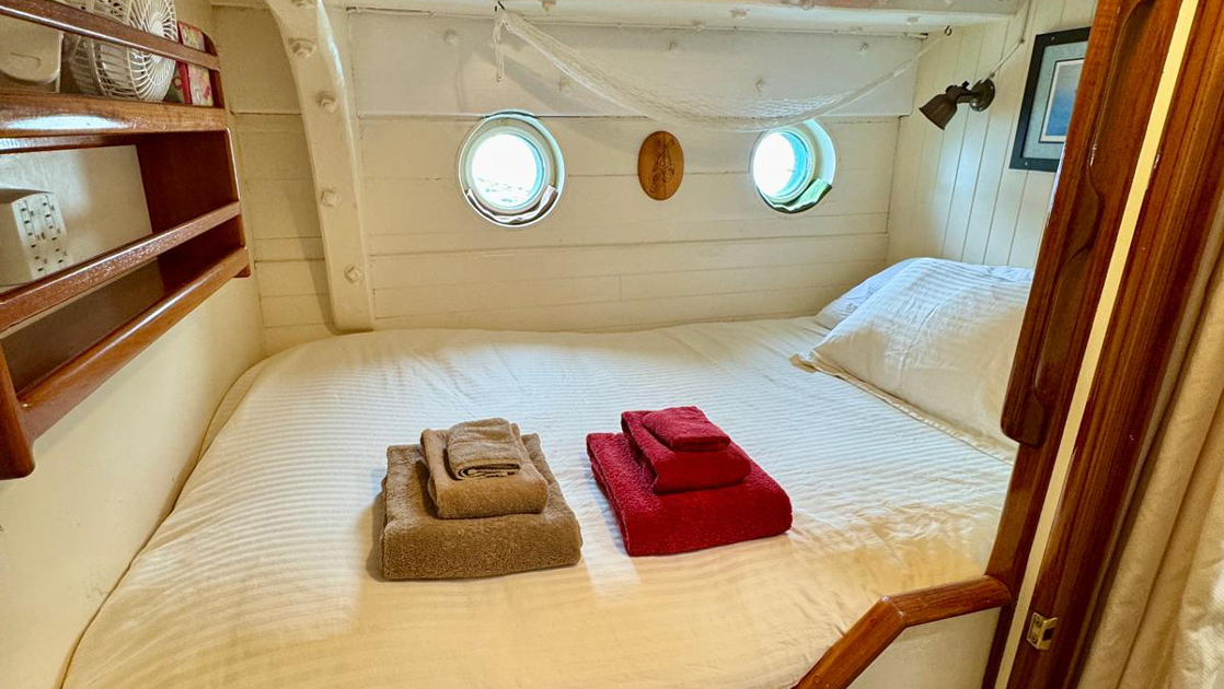 Cabin 2 on mv Catalyst with fixed wooden double bed in white linens with red & gold towels, portholes, white walls & bookshelf.