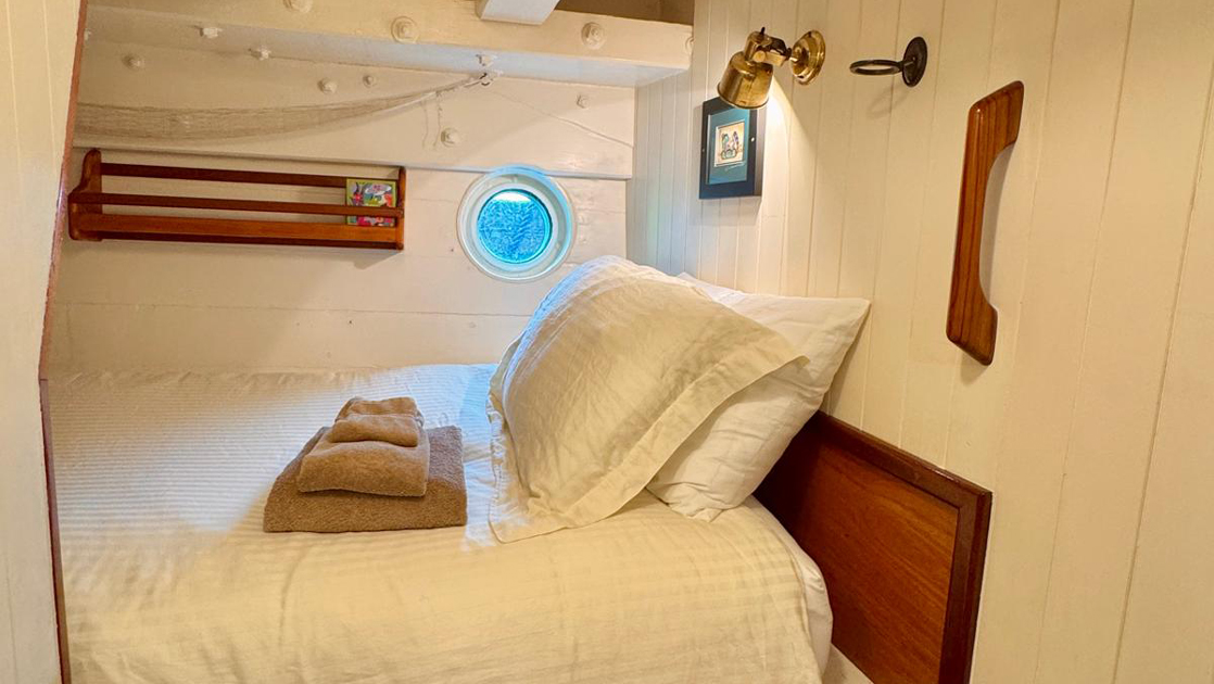 Cabin 4 on mv Catalyst with fixed wooden single bed in white linens with gold towels, porthole, white walls & bookshelf.