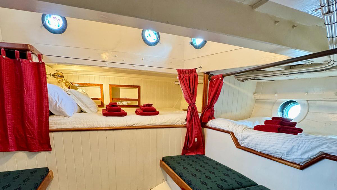 Cabin 5 on mv Catalyst with fixed double bed & 2 twin beds, all in white linens with red towels, portholes, white walls & green bench.