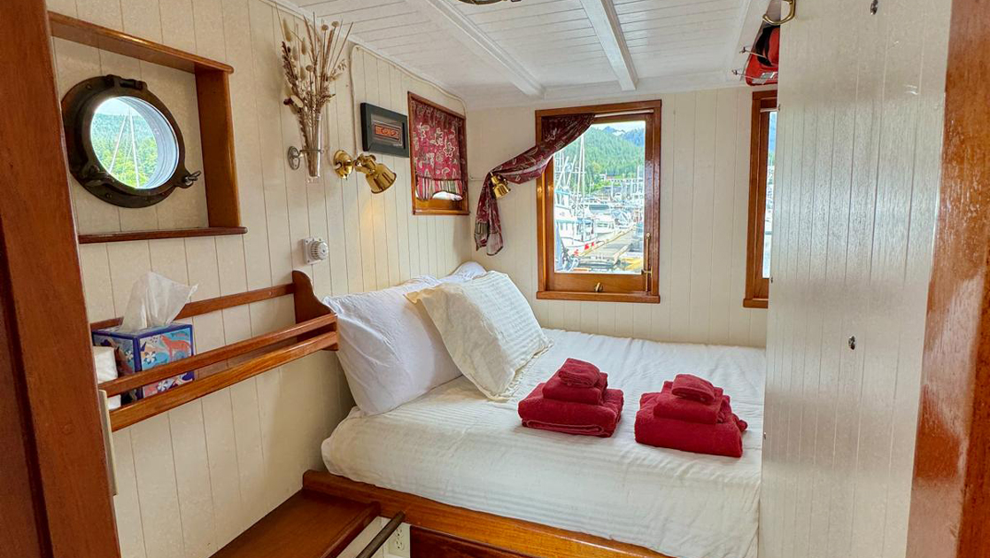 Cabin 6 on mv Catalyst with fixed wooden double bed in white linens with red towels, porthole & windows, white walls & bookshelf.