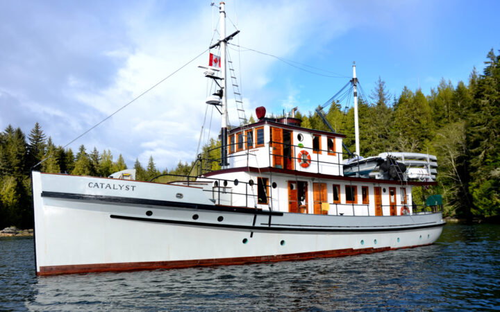 Port side of Catalyst historic wooden ship with white exteriors, square windows & tall mast under blue sky.