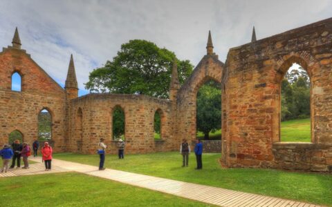 people stand on the grass in front of the stone walls of port arthur in australia on a cloudy day