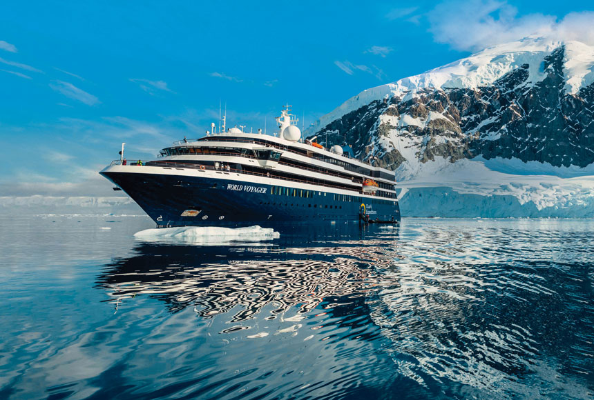 World Voyager cruise ship with blue hull & white upper decks sits in glassy Antarctic waters beside snowy peaks in the sun.