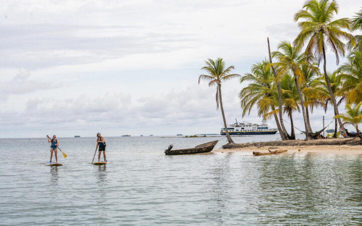 Man & woman paddle stand-up paddlebaords by shore of tiny island with white-sand beach & palm trees as ship sits nearby in Panama.