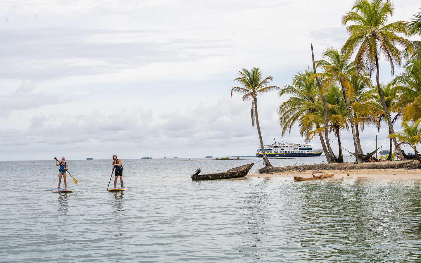 Man & woman paddle stand-up paddlebaords by shore of tiny island with white-sand beach & palm trees as ship sits nearby in Panama.