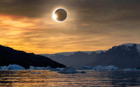 Solar eclipse reflects over cold icy waters with icebergs and dark rocky mountains.