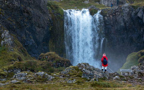 hiker in red standing near beautiful Waterfall with black rocks and green landscapes