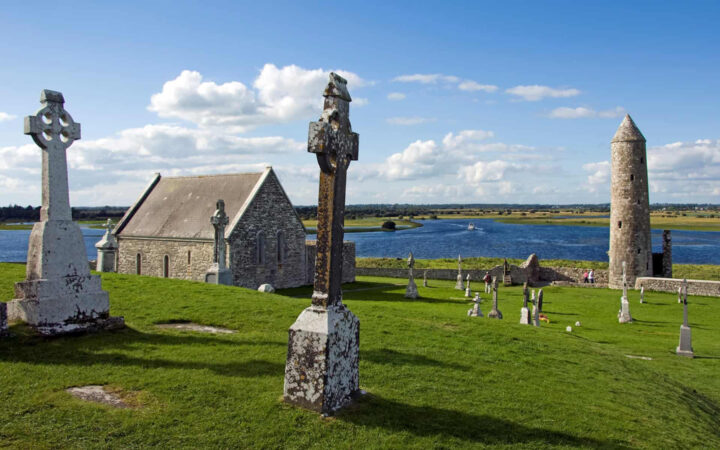 Small old stone church by green grass, stone crosses & calm river on a sunny day of the Classic Ireland River Cruise.