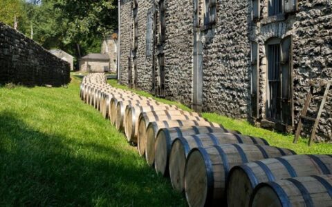 Line of whiskey barrels sits in a row along green grass & a stone building, seen during an Ireland barge cruise.