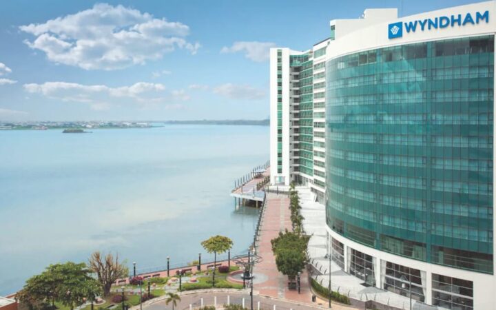 Exterior of Wyndham Guayaquil Hotel, multiple-story glass-encased building overlooking a large scenic river and a riverside walkway on a sunny day.
