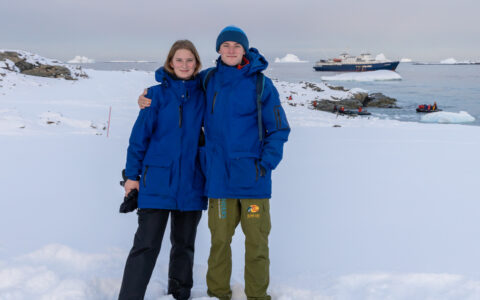 Young man & woman in blue jackets stand on snowfield by calm sea with a blue & white small ship in the distance in Antarctica.