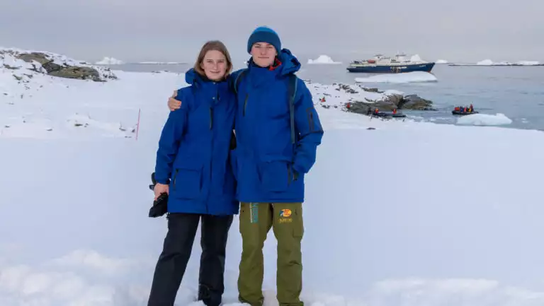 Young man & woman in blue jackets stand on snowfield by calm sea with a blue & white small ship in the distance in Antarctica.