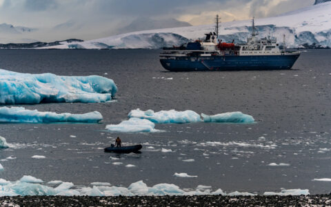 Small ship with blue hull & white upper decks sits in calm sea among blue icebergs as Zodiac cruises nearby in Antarctica.