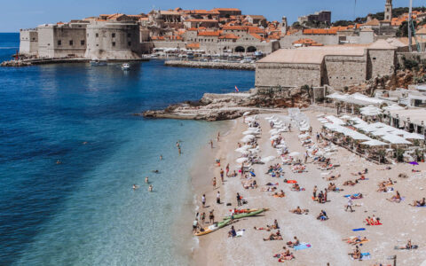 A beach scene in Dubrovnik Croatia seen from above with kayaks on the sandy shoreline and lots of white umbrellas