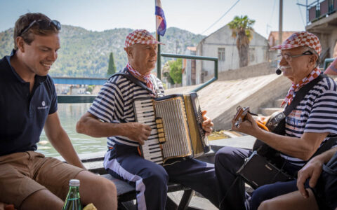 Two older men in blue and white striped shirts and checkered red hats play accordion and guitar with a younger man in a blue shirt and khaki shorts smiles beside them