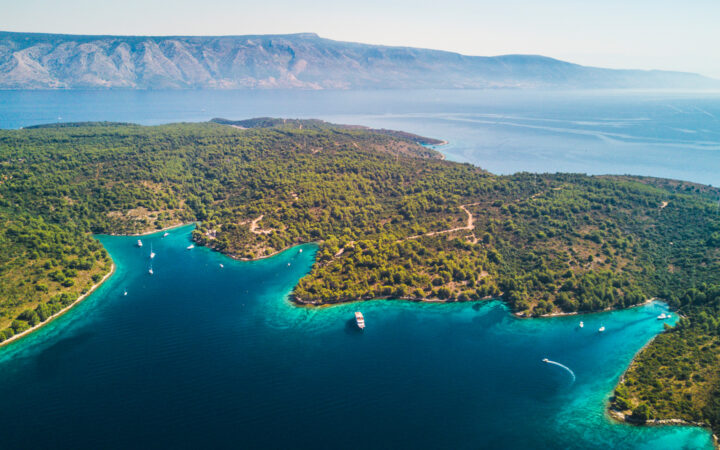 A small yacht is seen way down by the shore, among other small boats, on a remote shoreline in Croatia.