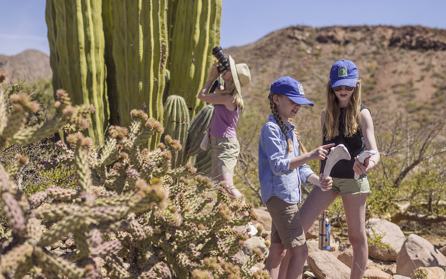 Two young girls point at an animal bone while their mother takes a picture of a cactus behind them