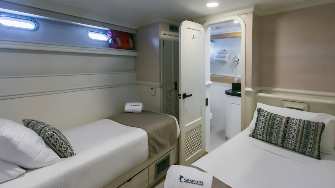Deluxe below-deck cabin with 2 twin beds on mv Galapagos Sky, with white wood accents & closet, bedside table, carpet & portholes.
